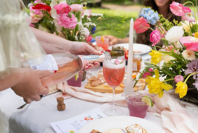 Hand pouring sparkling rosé into a frothy pink cocktail on a flower-filled outdoor brunch table with colorful spring blooms and appetizers.