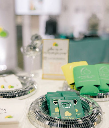 Event swag table with bright green and yellow promo bottle openers and can koozies neatly arranged on reflective trays, napkins visible in the foreground.