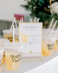 Gold-framed sign labeled “Signature Drink: Southern Charm” (sweet tea vodka and lemonade) surrounded by disposable plastic cups with lids and white straws filled with pale-yellow cocktails.