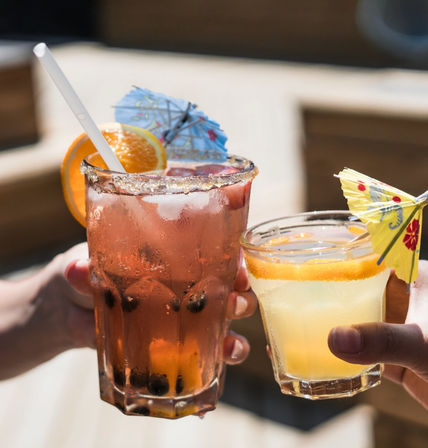 Two colorful summer cocktails clinking on a sunny outdoor patio — a tall pink, sugar-rimmed drink with orange slice, berries and blue umbrella, and a short citrus cocktail with orange peel and yellow umbrella.
