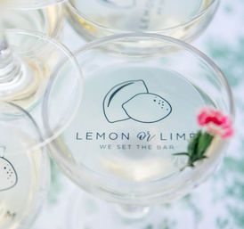 Close-up of coupe glasses with a minimalist lemon illustration printed on the glass, pale cocktails and a tiny pink flower garnish.