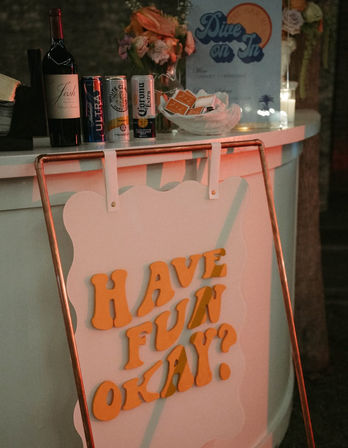Party bar counter with wine bottle, canned beers and seltzer, flowers and cards, playful retro sign on an easel reading “HAVE FUN OKAY?”