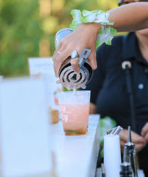Bartender pouring a pink tropical cocktail from a metal shaker through a strainer into an ice-filled plastic cup at an outdoor bar, wrist adorned with a green lei