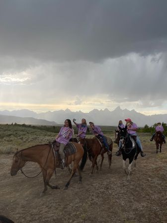 Riders in matching purple shirts on horseback crossing a sagebrush plain under dramatic storm clouds with a jagged mountain range and sunset light, one rider holding up a phone for a selfie.
