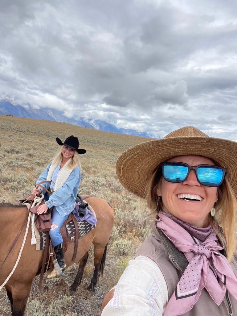 Selfie of two women horseback riding across a sagebrush prairie with distant mountains and dramatic cloudy sky — straw hat with mirrored sunglasses and a rider in a black cowboy hat on a brown horse.