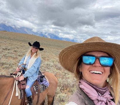 Selfie of two women horseback riding across a sagebrush prairie with distant mountains and dramatic cloudy sky — straw hat with mirrored sunglasses and a rider in a black cowboy hat on a brown horse.