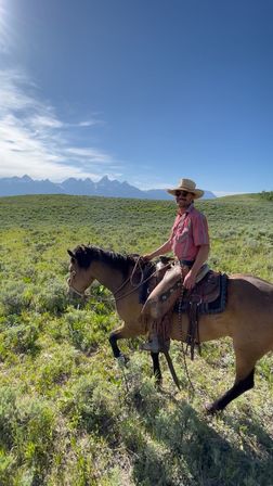 Cowboy on horseback crossing sagebrush prairie with distant jagged mountain peaks under a wide blue sky
