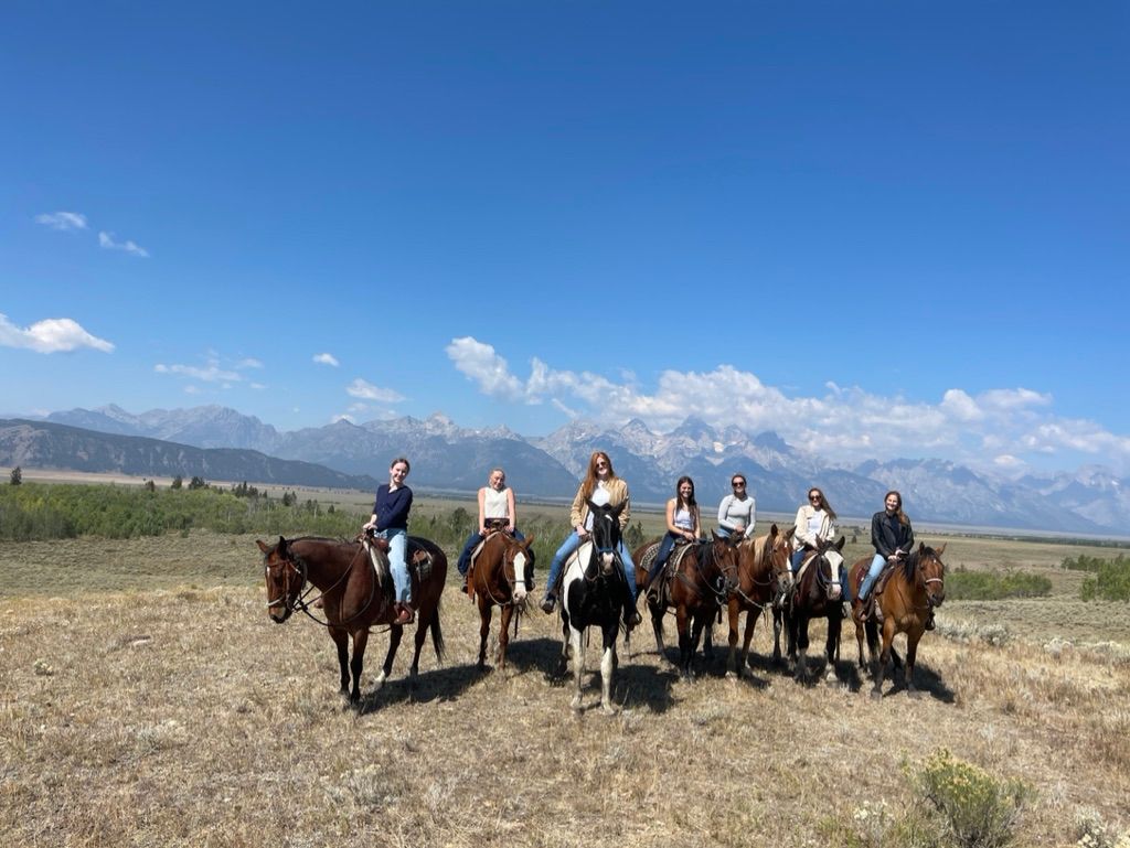 Eight riders on horseback crossing a dry sagebrush plain with a dramatic jagged mountain range and clear blue sky in the background — scenic western horseback ride