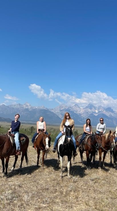 Eight riders on horseback crossing a dry sagebrush plain with a dramatic jagged mountain range and clear blue sky in the background — scenic western horseback ride