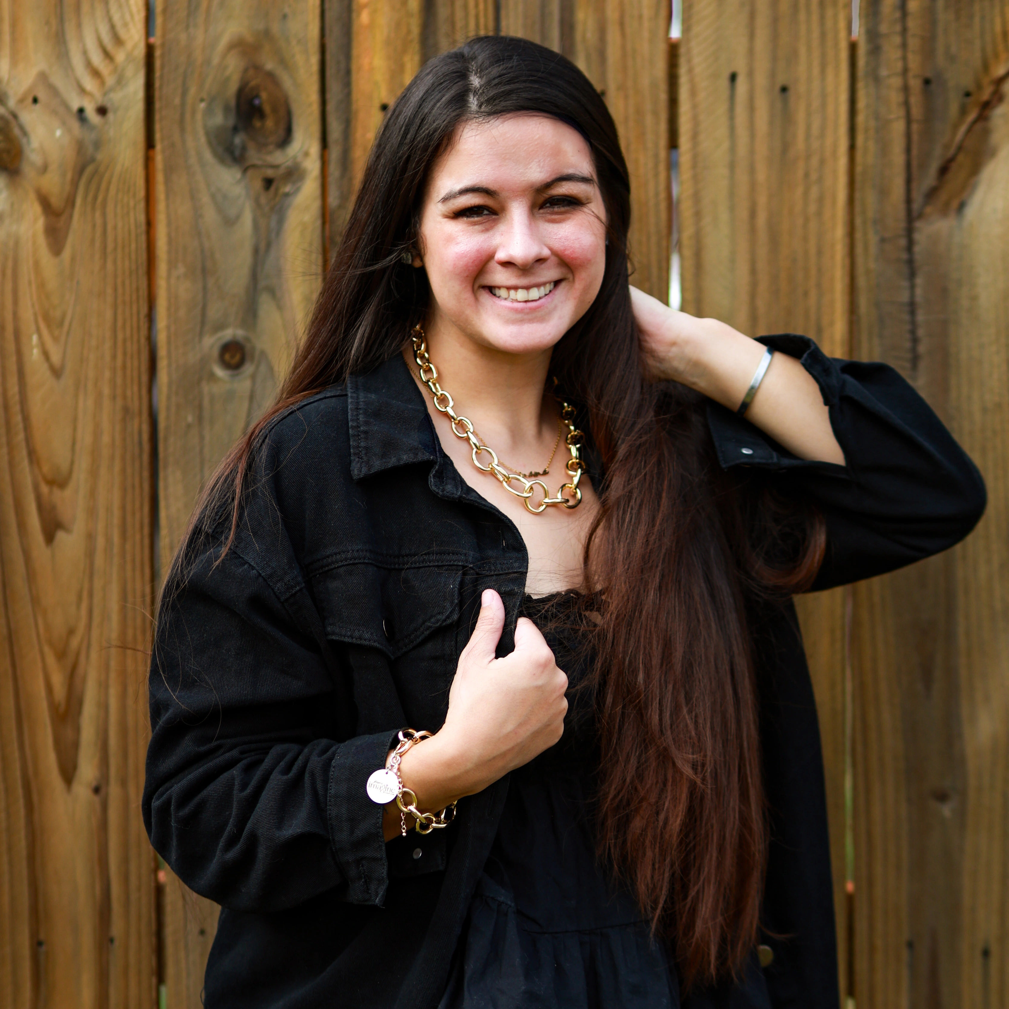 Outdoor portrait of a smiling woman with long brown hair in a black jacket, wearing a chunky gold chain necklace and bracelet, posed in front of a wooden fence.