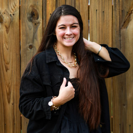 Outdoor portrait of a smiling woman with long brown hair in a black jacket, wearing a chunky gold chain necklace and bracelet, posed in front of a wooden fence.