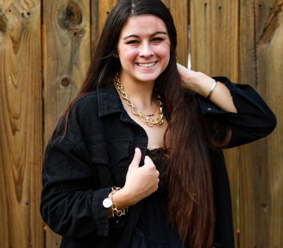 Outdoor portrait of a smiling woman with long brown hair in a black jacket, wearing a chunky gold chain necklace and bracelet, posed in front of a wooden fence.