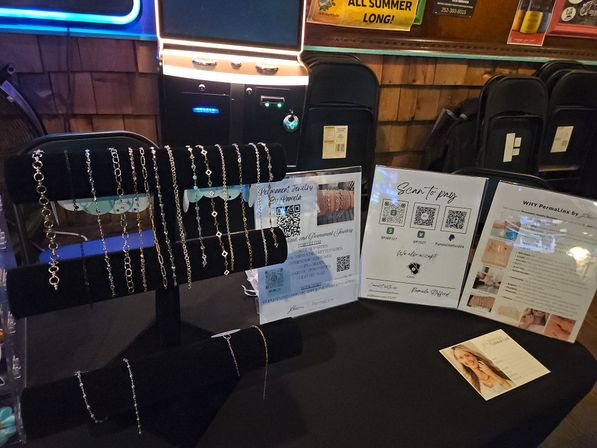 Vendor table at an indoor market with black velvet display racks holding metal chain bracelets, QR-code 'scan to pay' signage and printed brochures on a dark tablecloth.