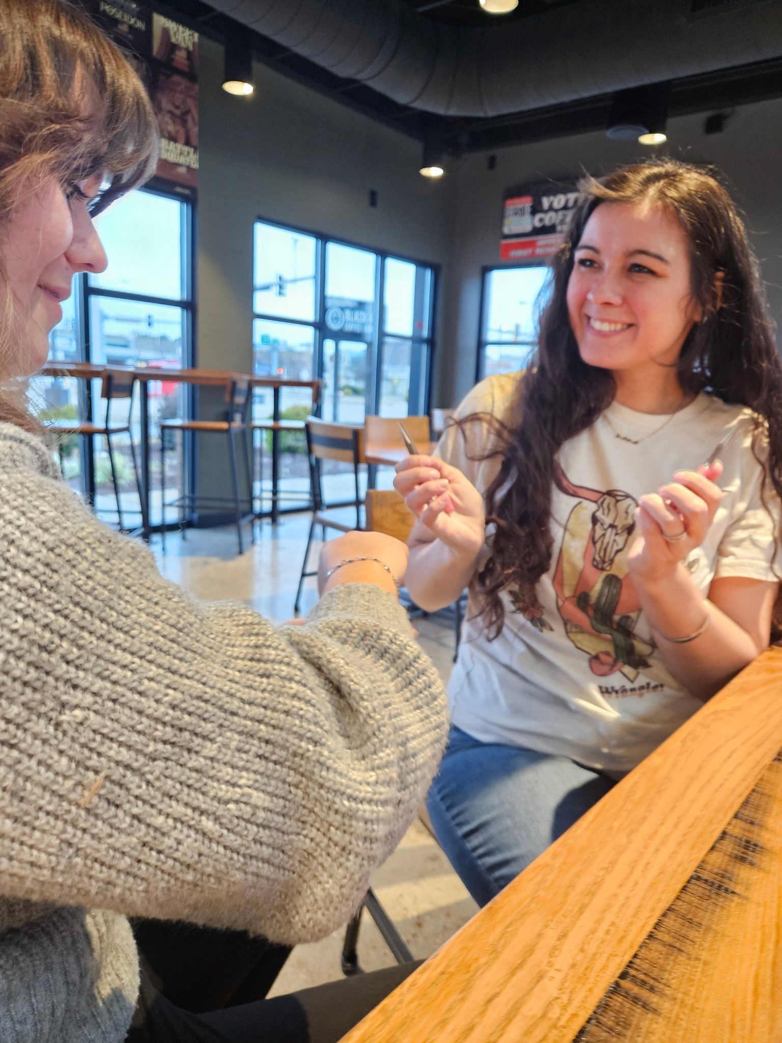 Two friends smiling across a wooden table in a bright modern coffee shop, holding small spoons and chatting by large windows.