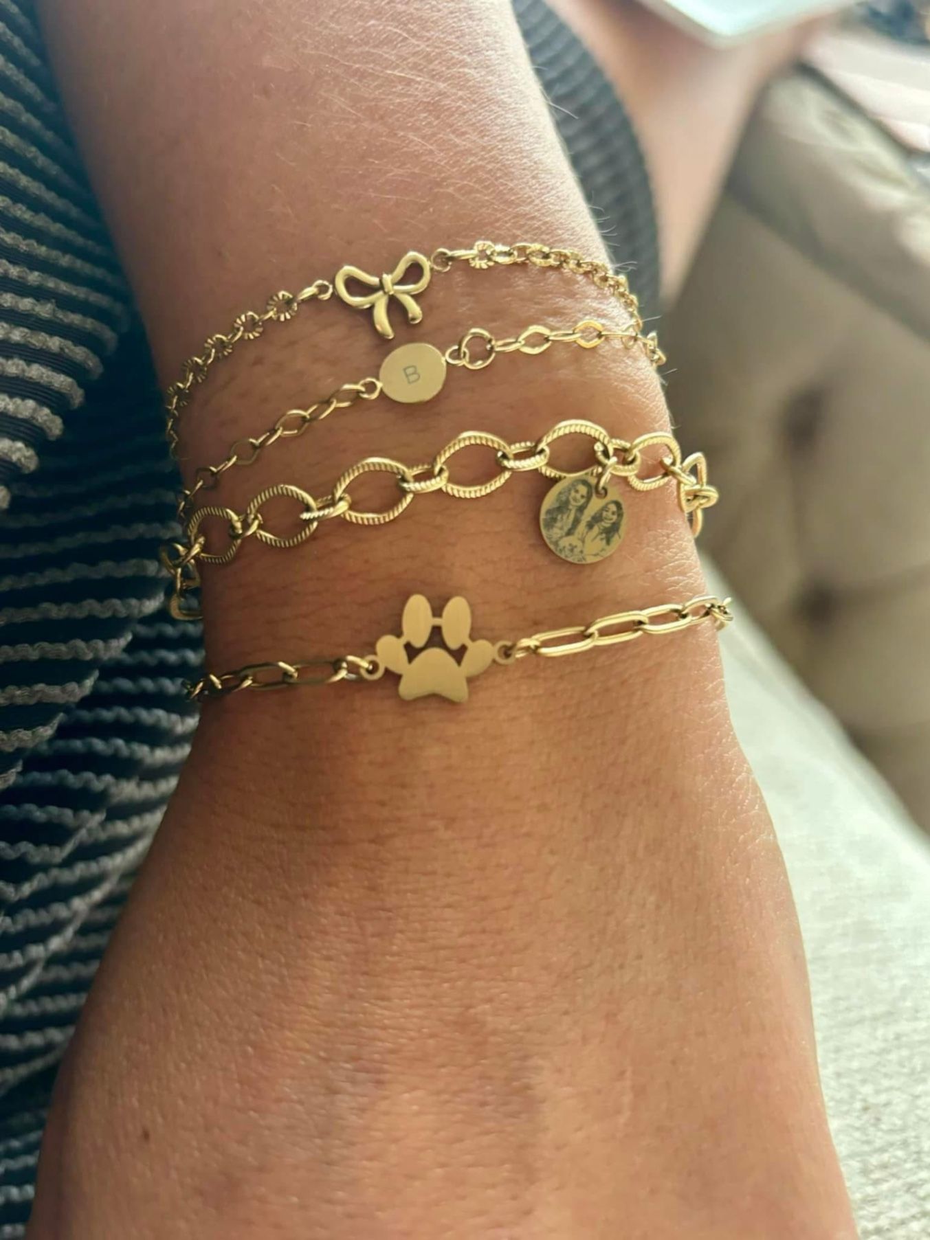 Close-up wrist shot of stacked gold charm bracelets: bow, 'B' initial disc, photo medallion and paw charm.