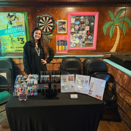 Person standing behind a black-draped vendor table displaying handmade jewelry, stickers, and flyers inside a wood-paneled bar with a dartboard, framed sticker collage, tropical palm mural, and beer promo poster.