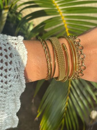 Sunlit stack of gold bracelets and bangles on a wrist with a white crochet sleeve, blurred tropical palm leaves in the background.