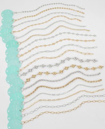 Flat-lay of gold and silver delicate chain bracelets in assorted link styles, arranged in parallel rows on a white background with teal circular product tags