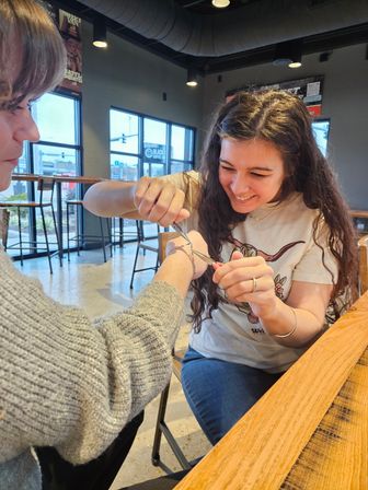 Smiling woman playfully uses small pliers to cut a bracelet from a friend’s wrist at a bright urban coffee shop table with large windows and bar stools in the background.