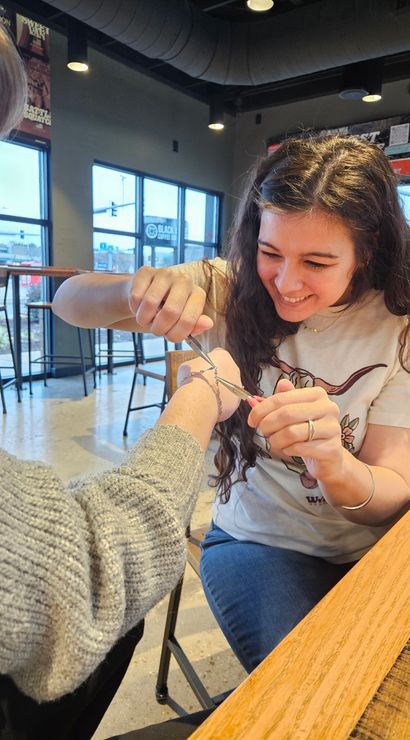 Smiling woman playfully uses small pliers to cut a bracelet from a friend’s wrist at a bright urban coffee shop table with large windows and bar stools in the background.