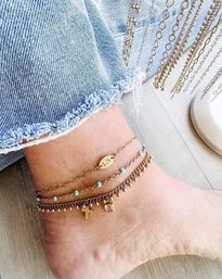 Close-up of ankle wearing layered gold anklets with turquoise beads, tiny cross and charm; frayed denim hem and hanging chain display on pale wood — boho summer style.
