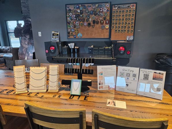Indoor artisan jewelry pop-up on a rustic wooden table—rows of necklaces and bracelets on cream and black display stands, informational signs with QR codes, and a corkboard of pins and patches on the gray wall behind; chairs and a lounge area with window visible to the left.