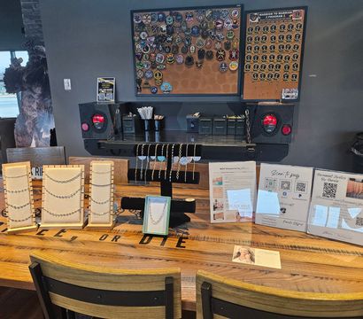 Indoor artisan jewelry pop-up on a rustic wooden table—rows of necklaces and bracelets on cream and black display stands, informational signs with QR codes, and a corkboard of pins and patches on the gray wall behind; chairs and a lounge area with window visible to the left.