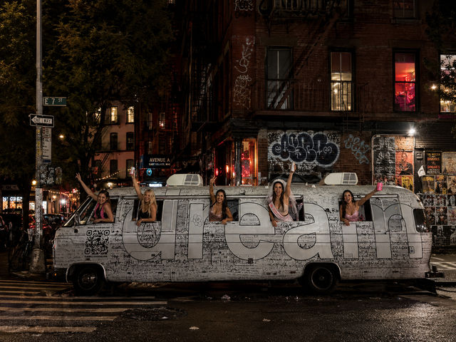 Five people lean from windows of a graffiti-covered white party van on a wet East Village, NYC street at night, waving drinks and celebrating under neon lights and brick buildings.