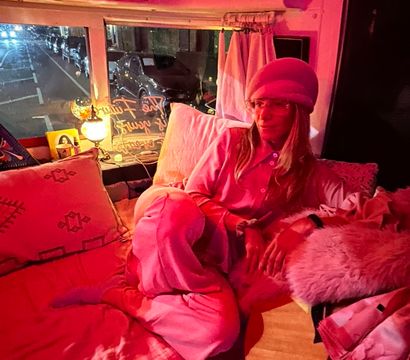 Cozy pink-lit campervan interior with a person in a pink beret lounging on pillows, acoustic guitar nearby, and a nighttime city street visible through the window.
