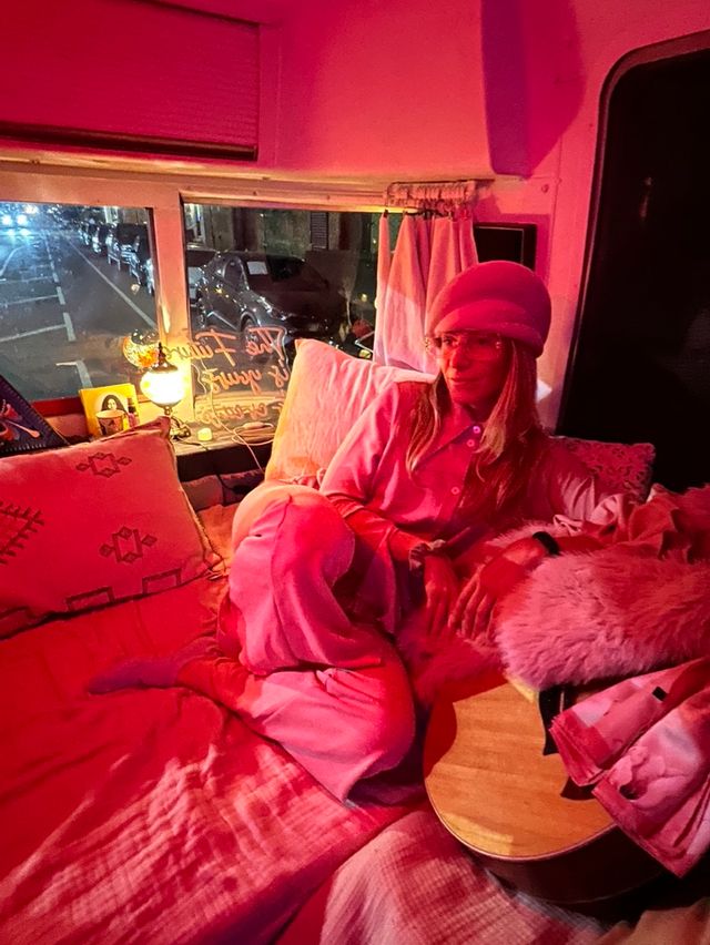 Cozy pink-lit campervan interior with a person in a pink beret lounging on pillows, acoustic guitar nearby, and a nighttime city street visible through the window.