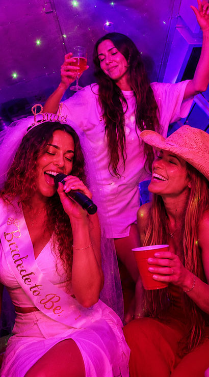 Bride-to-be singing karaoke on a neon-purple party bus, wearing veil and sash while friends in cowboy hats cheer with red cups and champagne.