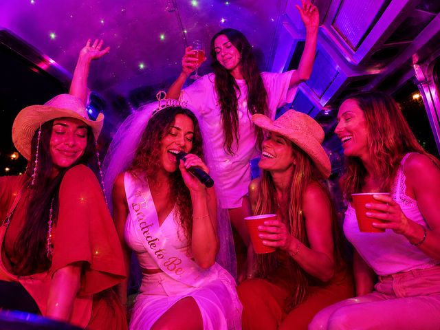 Bride-to-be singing karaoke on a neon-purple party bus, wearing veil and sash while friends in cowboy hats cheer with red cups and champagne.
