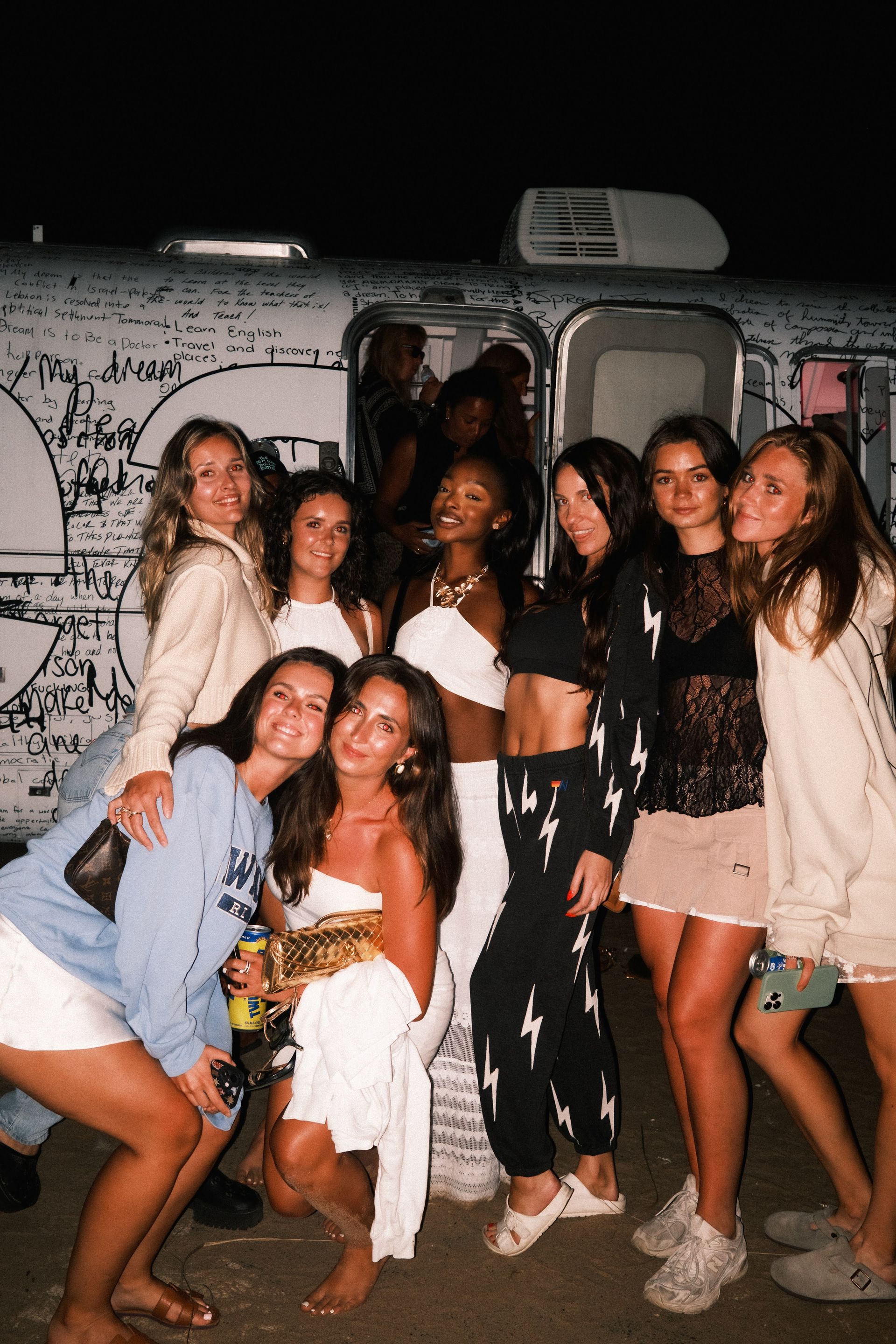 Group of smiling young women posing at night in front of a graffiti-covered silver trailer, wearing casual summer outfits, sandals and bare feet for a beach-party vibe.