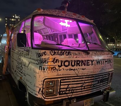 Graffiti-covered vintage camper van with handwritten messages and the word 'JOURNEY' on its front, glowing with magenta interior lights and parked on a downtown street at night.