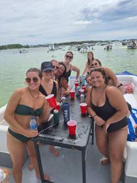 Smiling group of women in swimsuits at a pontoon boat party around a beer‑pong table with red cups, anchored boats and people in the shallow green coastal water