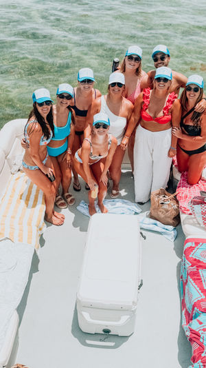 Smiling group of friends on a pontoon boat in shallow green coastal water, wearing matching blue trucker hats and summer swimwear on a sunny boat party.