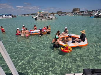 Crowded sandbar party in crystal-clear turquoise shallow water: groups in swimsuits gathered on inflatable platforms and pontoons playing drinking games with red cups, surrounded by anchored boats and party barges under a sunny blue sky.