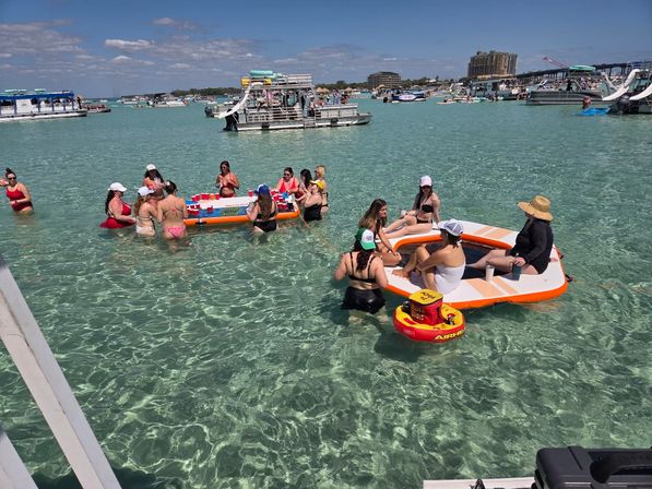 Crowded sandbar party in crystal-clear turquoise shallow water: groups in swimsuits gathered on inflatable platforms and pontoons playing drinking games with red cups, surrounded by anchored boats and party barges under a sunny blue sky.