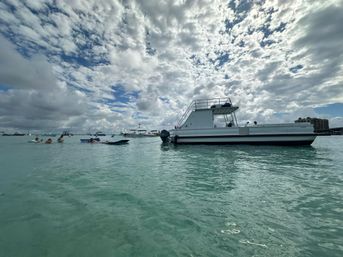 White double-decker pontoon boat anchored in clear turquoise bay with people on floats and many boats under a dramatic partly cloudy sky
