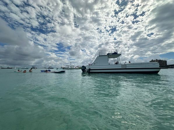 White pontoon boat anchored in clear turquoise shallow water with people on inflatables nearby under a dramatic puffy-cloud sky and distant coastal skyline