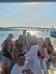 Smiling group of friends in swimsuits and celebratory sashes posing on a boat with sunlit water and a long coastal bridge at sunset in the background.