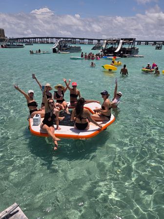 Group of people in swimsuits waving and relaxing on an orange-and-white inflatable platform in clear turquoise bay, surrounded by pontoon boats with water slides, inflatable tubes, and a low bridge under a sunny sky.