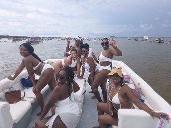 Seven women in white swimsuits smiling and posing on a lively boat party in a crowded bay, holding drinks and flashing peace signs under a cloudy sky.