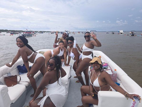 Seven women in white swimsuits smiling and posing on a pontoon boat, holding drinks and flashing peace signs, with dozens of boats and swimmers in a calm coastal bay under a cloudy summer sky.