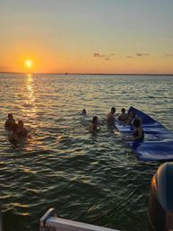 Group of people swimming and lounging on a blue inflatable mat beside a boat as the golden sun sets, casting a sparkling reflection across calm open water.