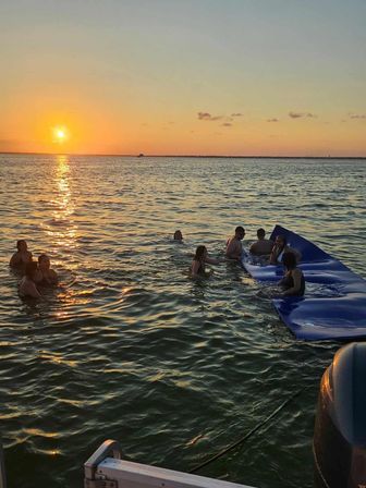 Golden sunset over open water with sun reflecting on ripples, a group of people swimming and lounging on a blue inflatable mat beside a boat.