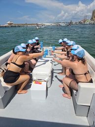 Group of friends wearing matching light-blue trucker hats toasting with drinks on a pontoon boat in sunny coastal waters with docks and sandy shoreline in the background.