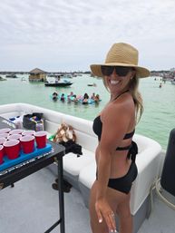 Smiling woman in a black bikini and straw sun hat on a party boat, red Solo cups on a table, friends on floaties and boats in a shallow turquoise bay with a tiki-style floating hut in the background