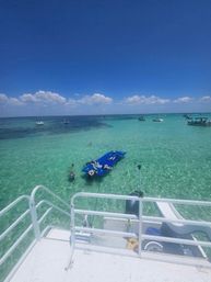 Sun-soaked boat deck overlooking crystal-clear turquoise shallow water, scattered boats on the horizon, and people lounging on a blue floating mat and wading beneath a bright blue sky with puffy clouds.
