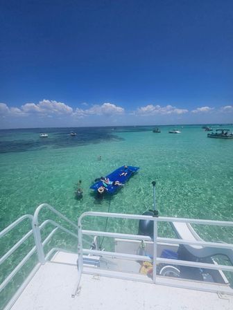 Overhead view of crystal-clear turquoise sea with anchored boats and people lounging on a blue floating mat beside a boat deck under a bright, cloud-dotted blue sky — tropical boating scene.